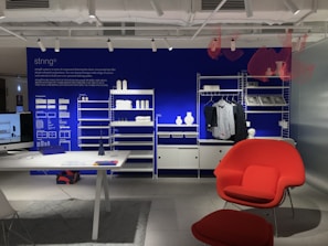 A modern interior space showcasing a minimalist shelving system against a vibrant blue wall. The shelves hold decorative items such as white vases and folded garments. A desk with a computer is positioned on the left side, while a bright red chair and ottoman set add a splash of color to the room. Industrial lighting fixtures hang from the ceiling.