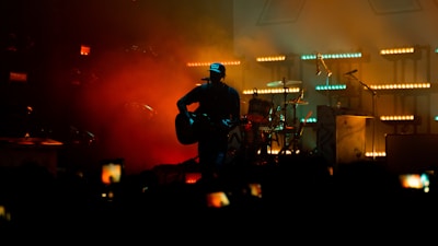 A musician stands on a stage playing a guitar. They are silhouetted against a backdrop of warm, colorful lights, including red, orange, and teal hues. A drum set and microphones are visible in the background, while several audience members are capturing the moment on their phones.