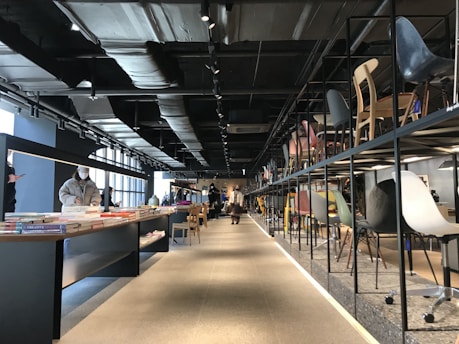 An interior space with modern industrial design, featuring a display of various colorful chairs on the right and books on the left. The ceiling is exposed with dark beams and ducts, contributing to an open and spacious atmosphere. There are people browsing through the items, some standing and one wearing a mask.