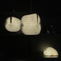 Two illuminated paper sculptures resembling fans or wings are arranged on a dark shelf. Their soft, white glow contrasts sharply with the black background, giving the appearance of floating light structures. A third similar object rests on the lower shelf, slightly smaller and with an open book-like shape.