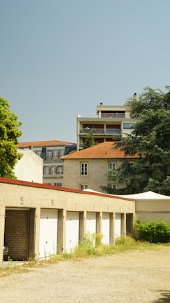 A series of numbered garage doors set against a backdrop of residential buildings and a clear blue sky. The garages have a concrete appearance with white doors, and some greenery is visible around them. In the background, there are two buildings: a mid-rise apartment complex with balconies and a cream-colored building with a red tile roof.