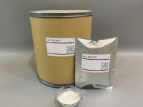A large cardboard cylinder and a silver foil bag, both labeled with 'CMOAPI' branding, are displayed. The labels include product details such as name, CAS number, weight, and a QR code. In front of them, a small glass dish contains a white powder with a metal spoon inside.
