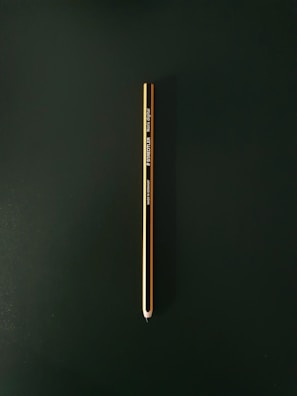 A single yellow and black pencil is placed vertically against a dark green background. The pencil is centered in the image and is sharpened to a point at the bottom. It features branding text and details printed along its side.