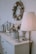 A stylish room corner features a decorated wreath on the wall, a table surface with a potted plant arrangement, metallic decor items, and a vintage-style rocking horse. A floor lamp with a pleated shade casts a warm glow over the setting.