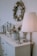 A stylish room corner features a decorated wreath on the wall, a table surface with a potted plant arrangement, metallic decor items, and a vintage-style rocking horse. A floor lamp with a pleated shade casts a warm glow over the setting.