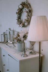 A stylish room corner features a decorated wreath on the wall, a table surface with a potted plant arrangement, metallic decor items, and a vintage-style rocking horse. A floor lamp with a pleated shade casts a warm glow over the setting.