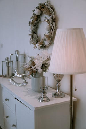 A stylish room corner features a decorated wreath on the wall, a table surface with a potted plant arrangement, metallic decor items, and a vintage-style rocking horse. A floor lamp with a pleated shade casts a warm glow over the setting.