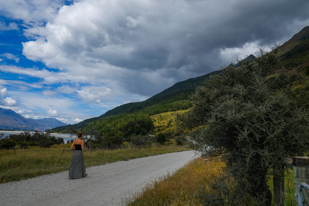 Mujer en vestido negro caminando en el camino entre el campo de hierba verde bajo nubes blancas durante el día