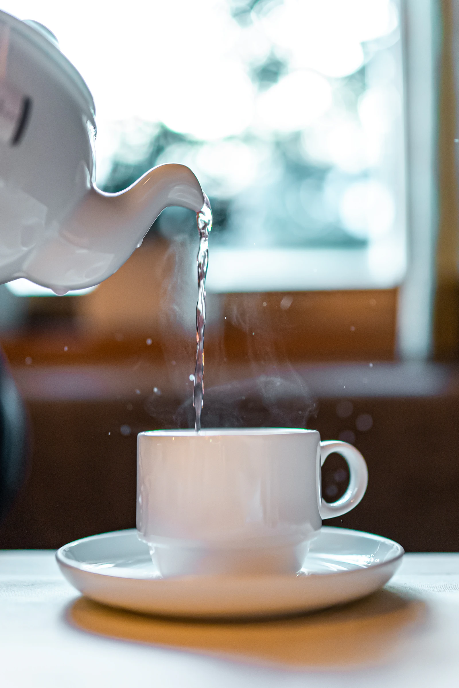 Tea being poured from a white ceramic pot into a cup, steam rising in warm morning light