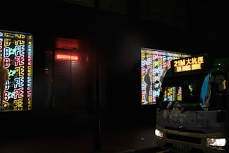 A bus is parked on the street at night, illuminated by neon signs featuring the word 'BAPE' in various colors. The bus's destination sign is lit up, displaying text in both English and Chinese. The atmosphere is urban and vibrant, with the bright colors of the neon contrasting against the dark surroundings.