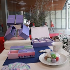 A display of pastel-colored gift boxes with elegant designs, likely for confectioneries or small goods. Various colors of macarons are arranged on a white plate, with a decorative cup nearby. The setup includes a table covered with a dark blue mat, brochures or cards, and a touch of floral decor. The background shows a modern, open space with large windows.