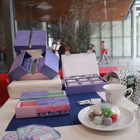 A display of pastel-colored gift boxes with elegant designs, likely for confectioneries or small goods. Various colors of macarons are arranged on a white plate, with a decorative cup nearby. The setup includes a table covered with a dark blue mat, brochures or cards, and a touch of floral decor. The background shows a modern, open space with large windows.