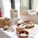 A tabletop display features various handmade and organic products, including garlic bulbs in baskets, loofah soaps, woven baskets, and natural packaging. The setting is bright with natural light coming through nearby windows.