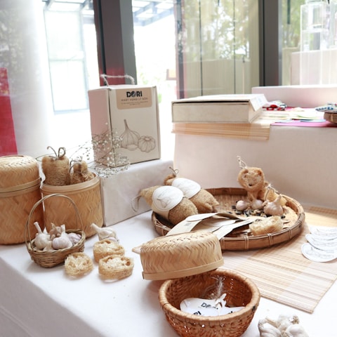 A tabletop display features various handmade and organic products, including garlic bulbs in baskets, loofah soaps, woven baskets, and natural packaging. The setting is bright with natural light coming through nearby windows.