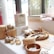 A tabletop display features various handmade and organic products, including garlic bulbs in baskets, loofah soaps, woven baskets, and natural packaging. The setting is bright with natural light coming through nearby windows.