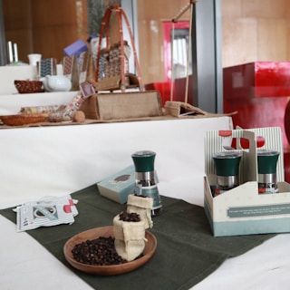 A wooden table displays various items, including a wooden bowl filled with black peppercorns, two small burlap sacks, a set of spice grinders in packaging, and some paper packets. There are decorative items and small containers arranged behind them against a backdrop of a red and brown interior.