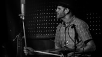 A drummer is captured playing in a dimly lit setting, likely a live music venue. The image is in black and white, focusing on the expressive, focused demeanor of the musician. The drummer is wearing a patterned shirt and a hat, and the drumsticks and microphone are prominently visible.