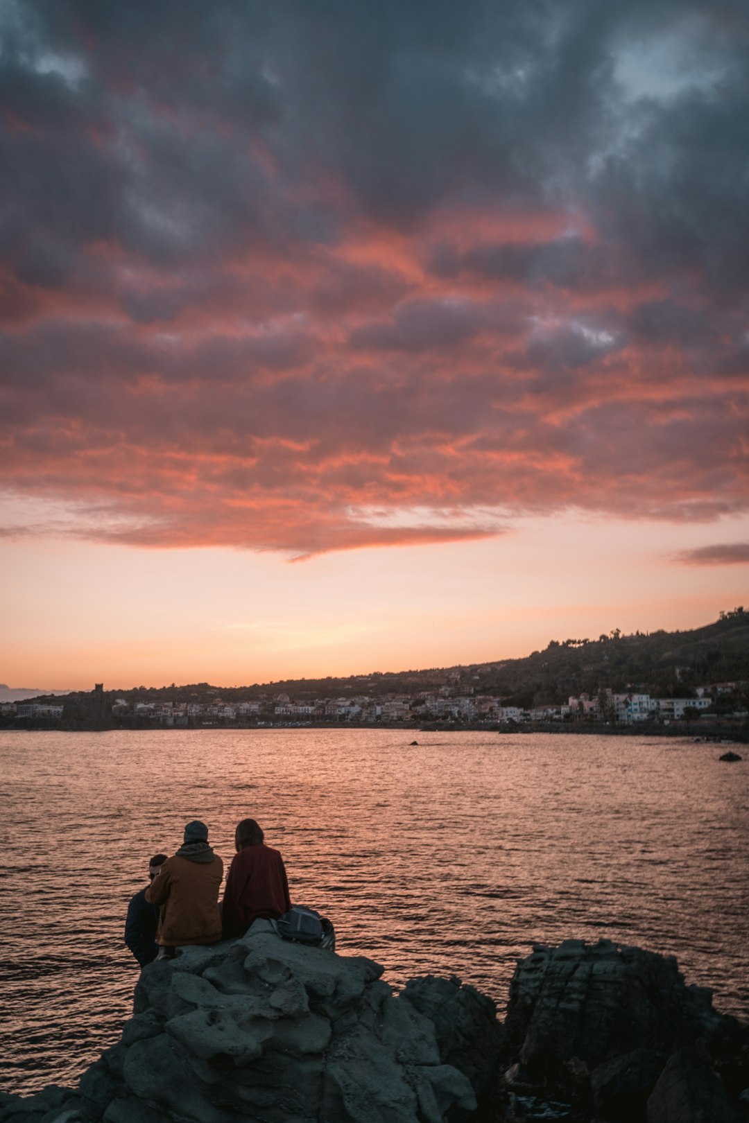 man and woman sitting on the ground looking at the sea during sunset