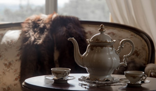 A vintage teapot and matching tea set are arranged on a wooden table. The teapot and teacups feature ornate designs with gold accents. They sit on a delicate lace doily. In the background, there is a floral-patterned sofa partially covered with a dark, fluffy throw. Soft lighting from a window creates a warm, inviting atmosphere.