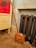 A wooden handled, brass bed warmer with a decorative perforated lid leans against an old cast iron radiator. The background includes a distressed blue wall and part of an antique cabinet with ornate detailing. The setting appears vintage and quaint.