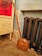 A wooden handled, brass bed warmer with a decorative perforated lid leans against an old cast iron radiator. The background includes a distressed blue wall and part of an antique cabinet with ornate detailing. The setting appears vintage and quaint.