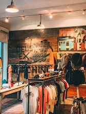A rustic store interior with wooden walls and stylish lighting. The wall features artistic graphics and text. Clothing items such as t-shirts and hats are neatly displayed on racks. The atmosphere feels laid-back and trendy.