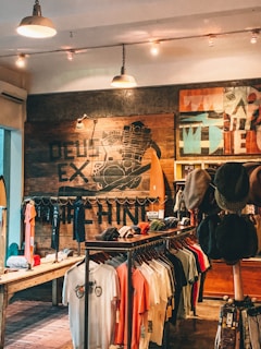 A rustic store interior with wooden walls and stylish lighting. The wall features artistic graphics and text. Clothing items such as t-shirts and hats are neatly displayed on racks. The atmosphere feels laid-back and trendy.