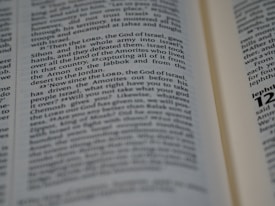 A close-up of an open book displaying text with varying font sizes, possibly a religious or reference book such as a Bible. The text is printed in black on cream-colored pages.