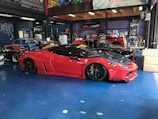 A sleek red sports car with black accents is parked inside a garage. The car is lowered and fitted with custom modifications, including black alloy wheels and aerodynamic enhancements. Surrounding the car are other vehicles and various automotive equipment, reflecting a vibrant and dynamic workshop environment.