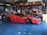 A sleek red sports car with black accents is parked inside a garage. The car is lowered and fitted with custom modifications, including black alloy wheels and aerodynamic enhancements. Surrounding the car are other vehicles and various automotive equipment, reflecting a vibrant and dynamic workshop environment.