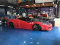 A sleek red sports car with black accents is parked inside a garage. The car is lowered and fitted with custom modifications, including black alloy wheels and aerodynamic enhancements. Surrounding the car are other vehicles and various automotive equipment, reflecting a vibrant and dynamic workshop environment.