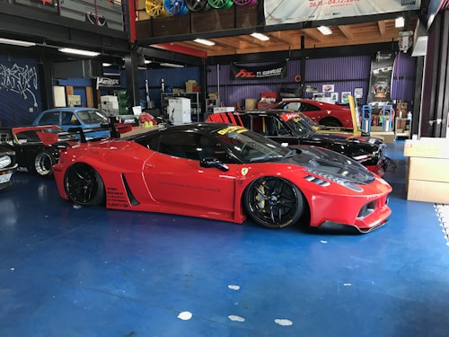 A sleek red sports car with black accents is parked inside a garage. The car is lowered and fitted with custom modifications, including black alloy wheels and aerodynamic enhancements. Surrounding the car are other vehicles and various automotive equipment, reflecting a vibrant and dynamic workshop environment.