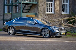 A sleek, black luxury sedan is parked outside a brick building with several white-paned windows and intertwined wooden branches. The vehicle's glossy surface reflects the surroundings, and its design includes large alloy wheels and a prominent front grille.