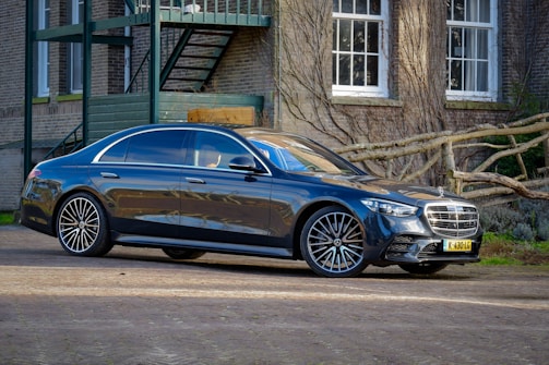 A sleek, black luxury sedan is parked outside a brick building with several white-paned windows and intertwined wooden branches. The vehicle's glossy surface reflects the surroundings, and its design includes large alloy wheels and a prominent front grille.