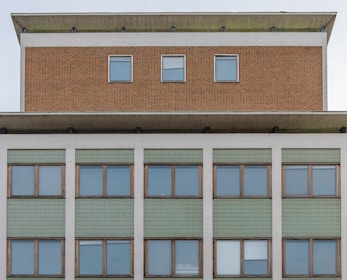 Facade of a multi-storey building with a symmetrical arrangement. The upper section features a brick texture and three evenly-spaced windows. Below, a larger section displays green tiling and numerous windows framed with wood.