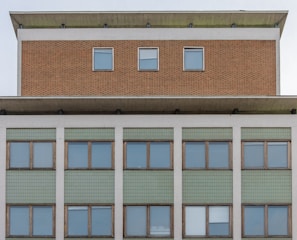 Facade of a multi-storey building with a symmetrical arrangement. The upper section features a brick texture and three evenly-spaced windows. Below, a larger section displays green tiling and numerous windows framed with wood.