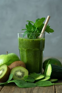 A green smoothie in a glass is surrounded by fresh ingredients, including whole and sliced kiwis, a cucumber, spinach leaves, and an apple. A straw and mint leaves garnish the drink, creating a refreshing and healthy presentation.