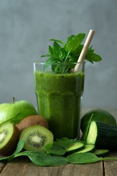 A green smoothie in a glass is surrounded by fresh ingredients, including whole and sliced kiwis, a cucumber, spinach leaves, and an apple. A straw and mint leaves garnish the drink, creating a refreshing and healthy presentation.