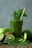 A green smoothie in a glass is surrounded by fresh ingredients, including whole and sliced kiwis, a cucumber, spinach leaves, and an apple. A straw and mint leaves garnish the drink, creating a refreshing and healthy presentation.