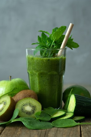A green smoothie in a glass is surrounded by fresh ingredients, including whole and sliced kiwis, a cucumber, spinach leaves, and an apple. A straw and mint leaves garnish the drink, creating a refreshing and healthy presentation.