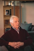 An elderly man with white hair sits in a cozy living room, dressed in a maroon sweater over a collared shirt. He appears introspective, with hands gently clasped in his lap. The background includes a wooden shelf with a framed photograph and a small ornament, contributing to a homely and serene atmosphere.