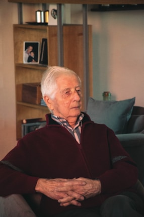 An elderly man with white hair sits in a cozy living room, dressed in a maroon sweater over a collared shirt. He appears introspective, with hands gently clasped in his lap. The background includes a wooden shelf with a framed photograph and a small ornament, contributing to a homely and serene atmosphere.