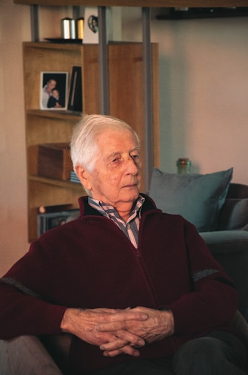 An elderly man with white hair sits in a cozy living room, dressed in a maroon sweater over a collared shirt. He appears introspective, with hands gently clasped in his lap. The background includes a wooden shelf with a framed photograph and a small ornament, contributing to a homely and serene atmosphere.