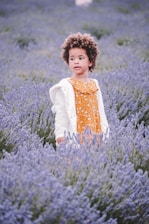 Illustration of Lírio, a cheerful child surrounded by soft lavender, green, and coral plants in a whimsical natural setting.