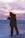 A silhouette of a person holding a camera, standing on a snowy surface with a backdrop of a serene sunset sky, displaying hues of purple, pink, and orange. The calm water in the distance reflects the soft colors of the sky.