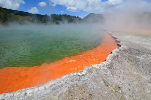 A geothermal hot spring with vibrant orange and green colors, surrounded by steam. The landscape includes rocky and mineral-rich terrain, with a mountainous backdrop and partially cloudy sky.