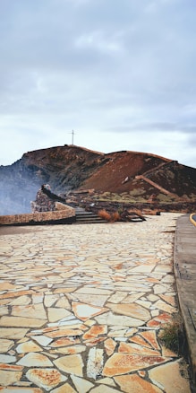 A rugged pathway with a tiled stone surface leads to a distant mountain peak. The mountain is topped with a large cross, visible against a cloudy sky. Sparse vegetation and rocky formations are present, with a person sitting on a stone structure in the distance.