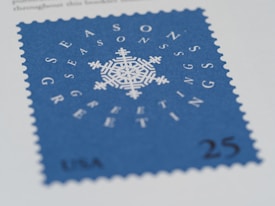 A blue postage stamp with a white intricate snowflake design in the center. Surrounding the snowflake are white letters spelling out 'SEASONS GREETINGS' in a circular pattern. The bottom of the stamp reads 'USA 25'. The edges of the stamp are serrated.