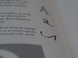 A close-up photograph of a book page featuring elegant swash capital letters, specifically focusing on the letter 'A'. The text is printed in a serif font, creating a classic and artistic look. The background is blurred, emphasizing the sharpness and detail of the letters.