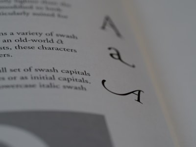 A close-up photograph of a book page featuring elegant swash capital letters, specifically focusing on the letter 'A'. The text is printed in a serif font, creating a classic and artistic look. The background is blurred, emphasizing the sharpness and detail of the letters.